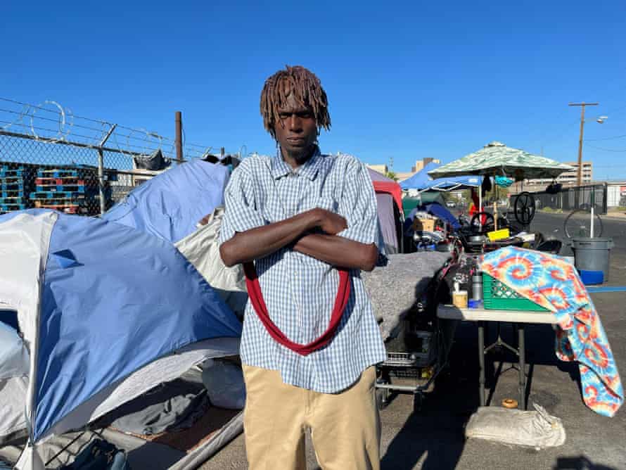 A man with his arms crossed stands in a homeless encampment that offers little shade.