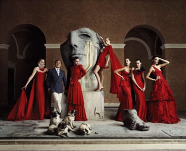 Valentino stands next to a sculture of a face surrounded by models in red dresses and four pugs