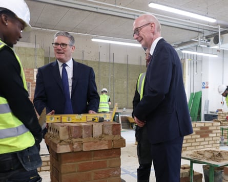 Starmer and McFadden talking to a person wearing a hi-vis gilet and a white hard hat working on a brick structure
