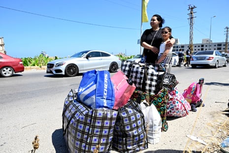A woman has her arm wrapped around her young daughter as they stand next to a large pile of bags under a blue sunny sky.