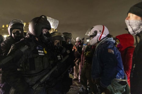 Against a dark sky, a line of federal agents in riot gear face off with a group of protesters bundled up in winter clothing. Some protesters are wearing helmets and face coverings.