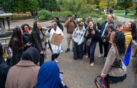 A group of about 20 young people of diverse ethnicities, including women in headscarves, stand together in a park and scream; they look happy and as if they are enjoying themselves; some are filming their scream. They are dressed warmly in coats and scarves.
