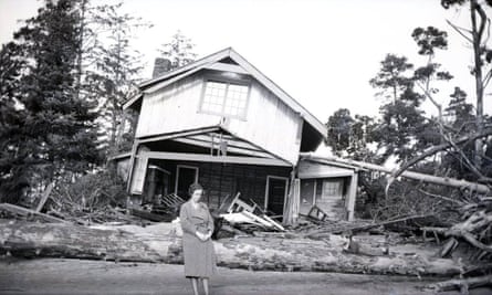 A woman stands in front of a house that has canted sideways, its supporting walls and beams destroyed.