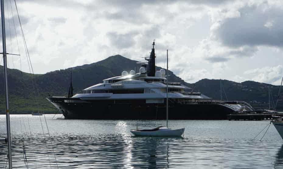 A sleek, black-hulled superyacht at anchor in a bay ringed by hills, photographed from water level, with a small sailing dinghy in the foreground