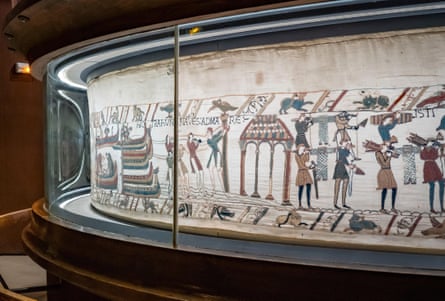 Tapestry on display behind glass in the showcase; a section showing boats landing and men marching is seen.