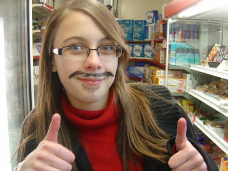Amelia Tait standing in a shop doing a double thumbs up, sporting a black, drawn-on moustache and beard, and wearing glasses, a red polo jumper and a brown blazer