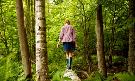 A woman in wellingtons walking along a fallen tree trunk in woodland.