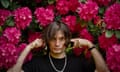 Press shot of Jonny Greenwood, he has his fingers in his ears. Behind him are large pink/purple flowers on a bush.