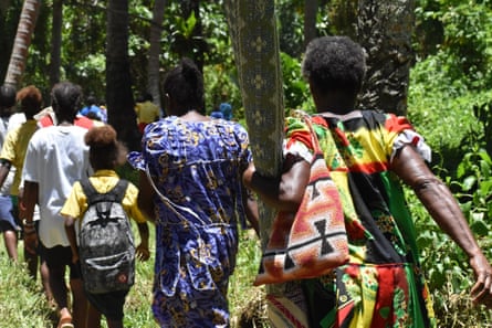 A line of people walk through a forest carrying bags and a small number of posessions.