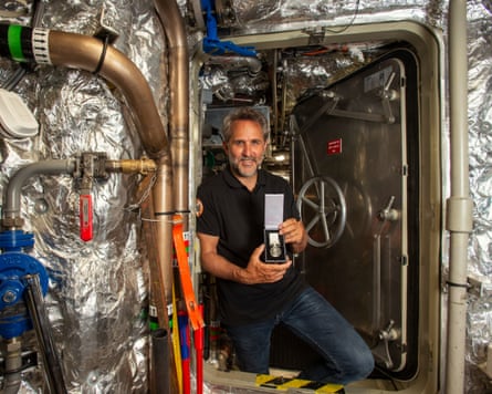 A bearded older man poses with a medal in a bulkhead door of a chamber lined with padded silver foil and pipes