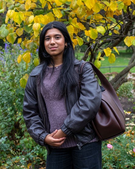 she has long, straight black hair and wears a black leather jacket and light mauve jumper; she carries a large brown leather bag and stands in front of a tree with yellow leaves.