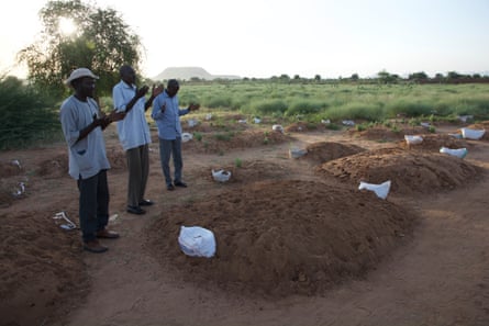 Three people stand next to fresh graves dug next to a grassy plain.