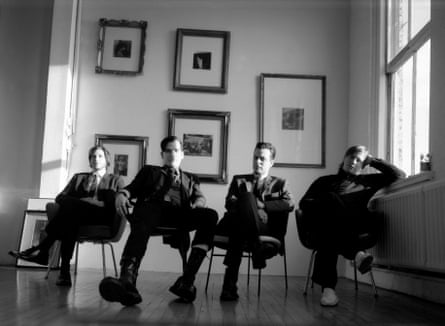 A black and white image of four men in dark suits sitting on chairs, with framed portraits behind them