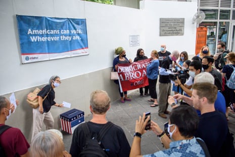 a woman drops a ballot in a box under a banner that reads 'Americans can vote wherever they are'