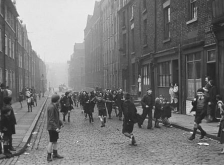 A large group of children play on a cobbled street with brick housing on either side