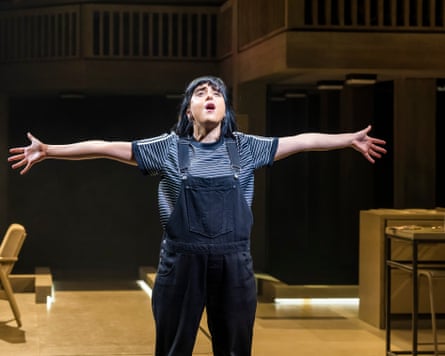 Memon, dressed in a blue striped T-shirt and dark blue dungarees, stands singing, arms outstretched