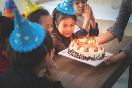 Cute little girl in party hat looks on as a cake with candles is put down before her