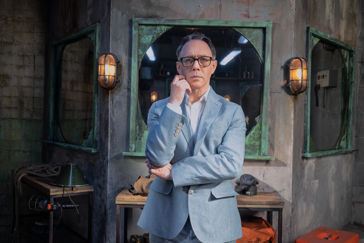Reece Shearsmith poses thoughtfully, arms folded, in a dim industrial room