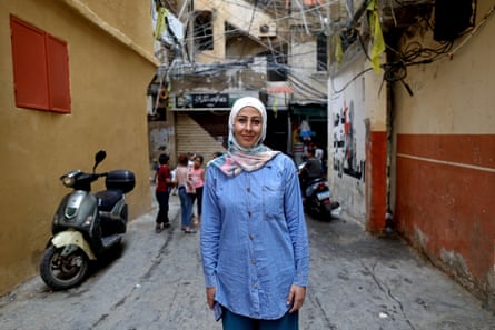 Batool Ghanem stands in a narrow street in Shatila, wearing a blue shirt and white headdress, with a moped behind her.