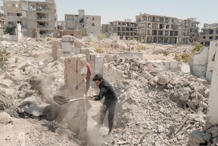 A man wields a spade amid a pile of rubble; damaged apartment blocks are in the background.