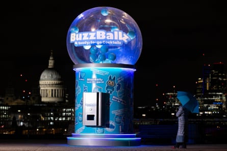 A giant blue BuzzBallz dispensing machine with London skyline in background and person holding an umbrella staring up at it
