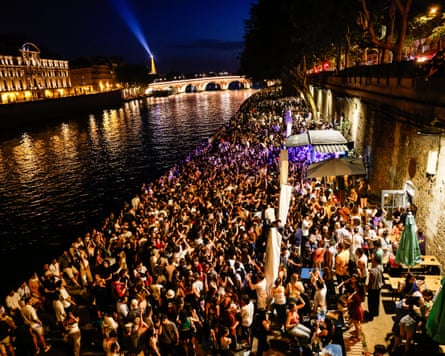 Large crowds gather on the walkway beside the Seine in Paris at night