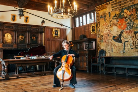 Woodcock playing the cello in a large room with wooden floor, wood panels and a large artwork on the wall