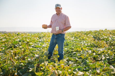 Smiling white man in a low green field wearing a ball cap, jeans, and short-sleeved collared shirt, holding what looks like a small pumpkin in one man and maybe a Starbucks cup in the other.