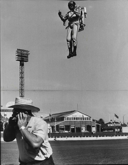 Black and white photograph showing Bob Courter  350ft above a showground ring using a jet pack strapped to his back. In front of him a man turns away holding his hand to the side of his head.