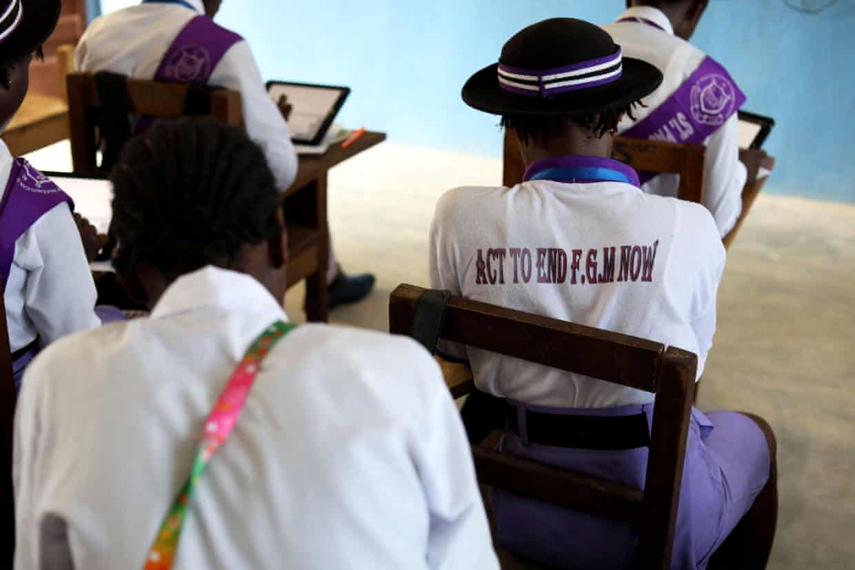Teenage girls sitting on chairs in a classroom, facing away from the camera. One wears a shirt with ‘Act to end FGM now’ written on the back.
