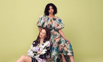 Two female models sit together, with dour expressions. One has long brown hair and wears a black, white and lilac print dress. The other has brown coils and wears a voluminous dress with a multicoloured print.