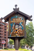 Sign reading Ye Olde Swiss Cottage with a brick building, tree and tube sign behind it