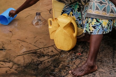 A woman reaches into cloudy water with a blue jug in order to fill two yellow water containers at her feet.