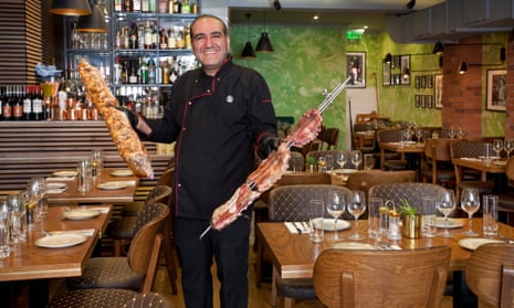 A smiling man stands in a dining room while holding meat on rotisserie skewers