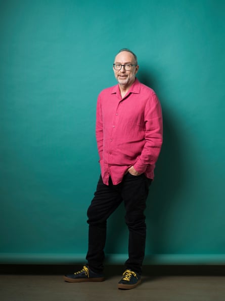 Full-length portrait of Jimmy Wales in pink shirt and black trousers against a blue/green backdrop