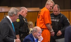 Man in orange jumpsuit stands in court with court officials while lawyers sit