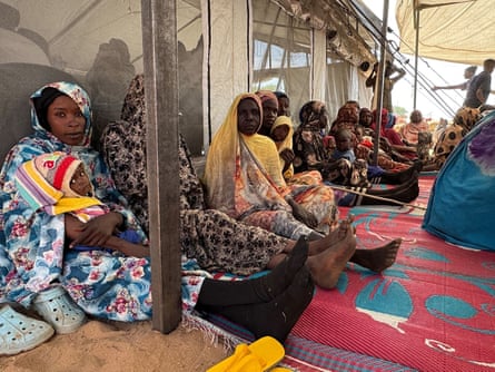 Women sit in lines under an awning next to a large tent.
