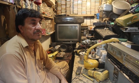 Man sitting in a tiny shop crowded with a TV and cassette players and music cassettes on shelves lining the walls.