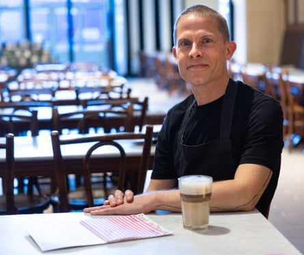 A man in a dark-blue T-shirt with a glass of coffee in front of him sitting at a table