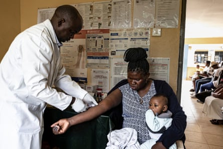 Taking a blood sample from a mother (holding a baby) living with HIV in Kisumu, Kenya.