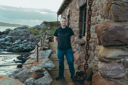 A middle-aged man stands outside a stone building by the sea.