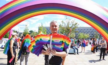 Robert Biedroń holds an armful of rainbow flags while standing in front of a giant rainbow arch