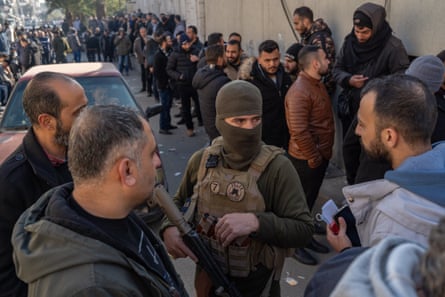 An HTS soldier wearing a balaclava manages a large crowd of former personnel from the Assad regime