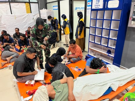 People sit on mats or lay on fold-up beds in a room, as officials and medical staff stand around