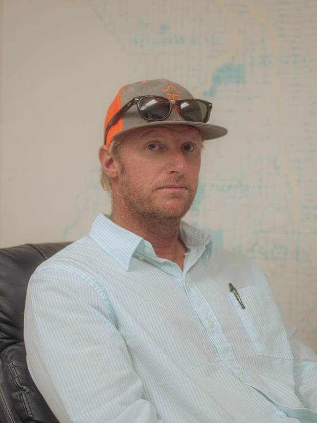 Man in a light blue shirt, brown and orange cap with sunglasses perched on top, looking into the camera