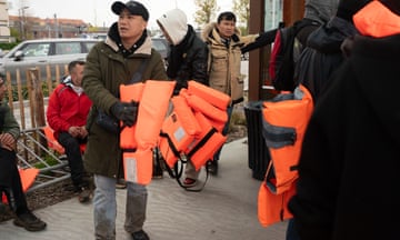 Man holds number of orange life jackets with others stood around next to bus
