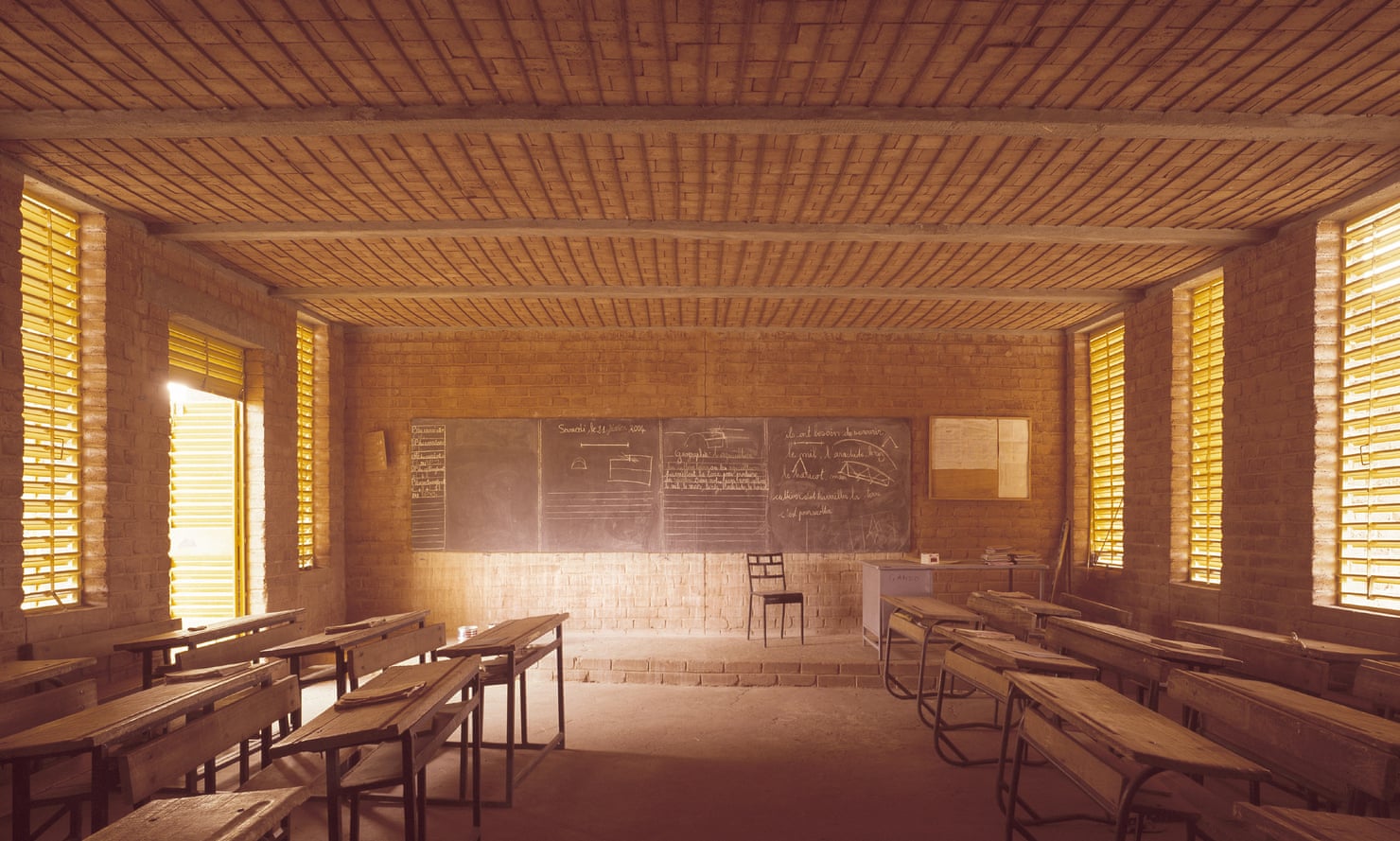 A classroom in Gando primary school, with red brick walls, wooden slats on the windows, wooden benches and a blackboard.