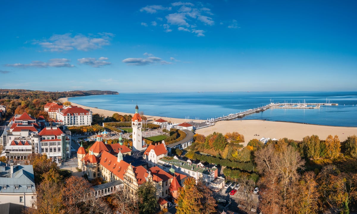 Overhead shot of a small town of whitewashed red-roofed houses, surrounded by trees and fringed by a sandy beach and blue sea