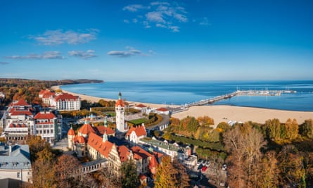 Overhead shot of a small town of whitewashed red-roofed houses, surrounded by trees and fringed by a sandy beach and blue sea