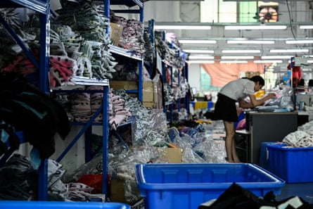 A woman stands bent over a table working in a factory setting. Around her are piles of clothes wrapped in plastic. 